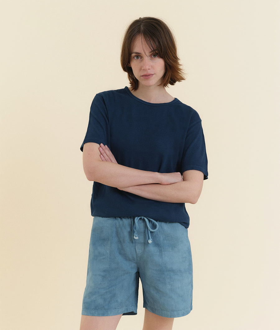The image shows a person wearing a navy blue t-shirt and light blue drawstring shorts standing with their arms crossed against a cream background - Industry of All Nations