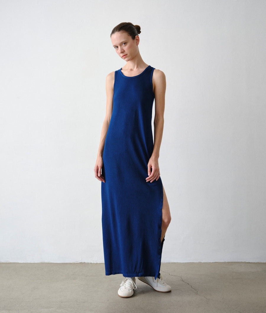 A woman models the IOAN Pique Tank Dress in Indigo 12, standing against a light gray studio wall and concrete floor.