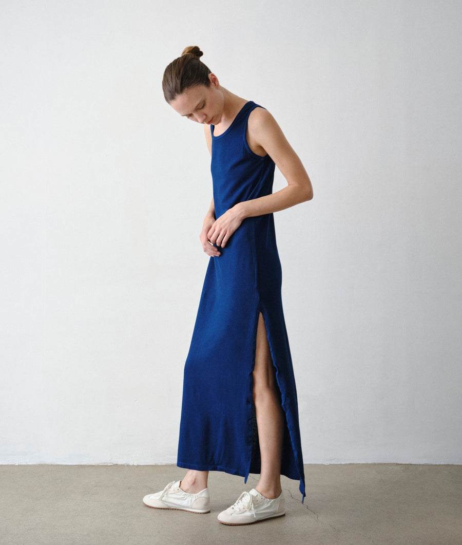 A side view of a woman modeling the IOAN Pique Tank Dress in Indigo 12, highlighting the side slit, against a light gray studio wall and concrete floor.