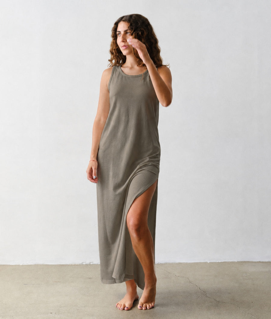 A woman models the IOAN Pique Tank Dress in Iron 4, a sleeveless, floor-length maxi dress in a muted green-gray tone with a high side slit, standing against an off-white studio wall and concrete floor.