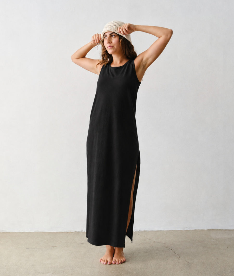 A woman models the IOAN Pique Tank Dress in Iron 8, a sleeveless black maxi dress with a high side slit, paired with a beige knit hat, standing barefoot against a light gray studio wall and concrete floor.