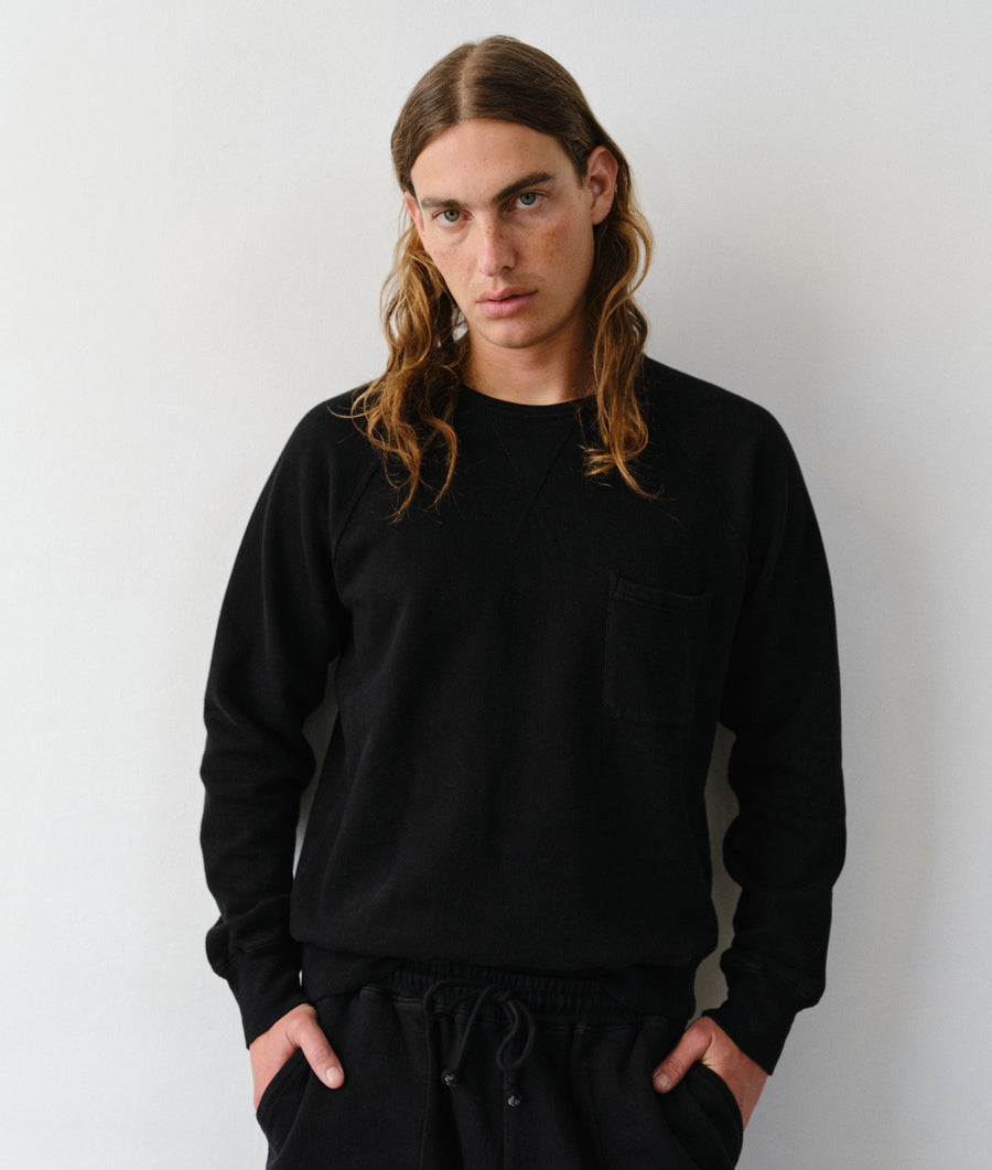 A man with long light brown hair wearing a black crewneck sweatshirt with a chest pocket and black sweatpants, standing against a plain light gray wall.