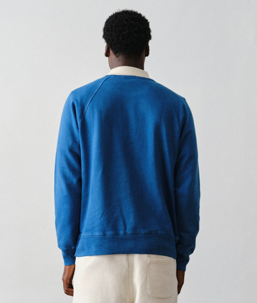 Back view of a man wearing the IOAN Pocket Sweatshirt in deep indigo blue and IOAN Super Sweatpants in cream against a neutral light gray background.