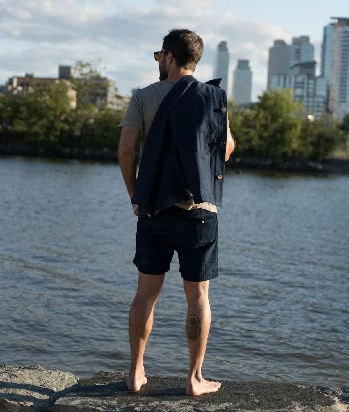 A man stands barefoot on a rock by a river, viewed from behind, wearing an IOAN Crewneck T-Shirt and Indigo 12 dark navy shorts, with a dark navy jacket draped over his shoulder. A city skyline and trees are visible across the water in the background.