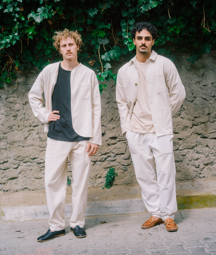 Two men standing outdoors in front of a textured stone wall with green ivy, wearing matching light beige linen-look outfits over layered neutral shirts and trousers.