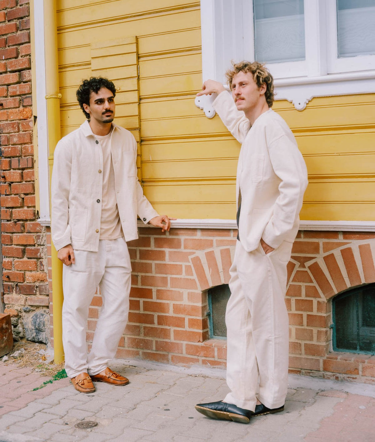 Two men wearing IOAN Ponya Corduroy Pants pose outdoors against a yellow wooden wall with a brick foundation.