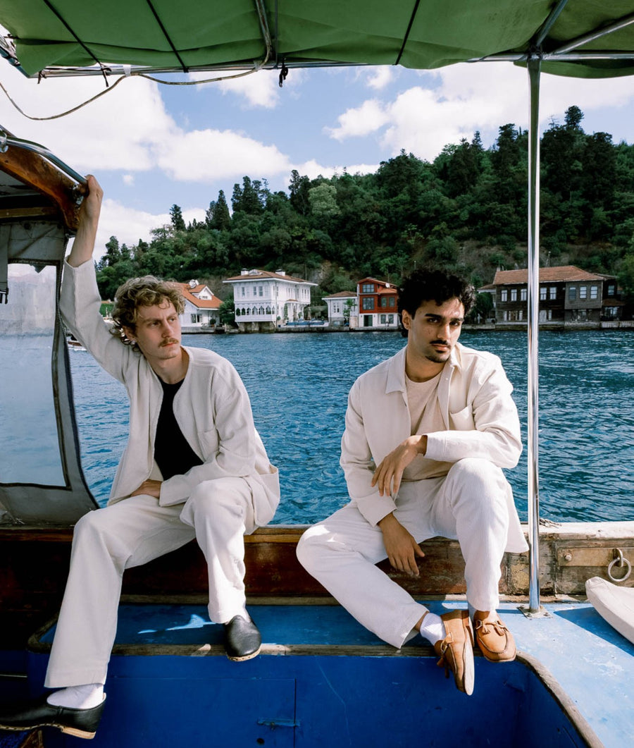 Two men in matching light cream suits sit on a blue boat under a green canopy, with a scenic lake and tree-covered shoreline in the background.