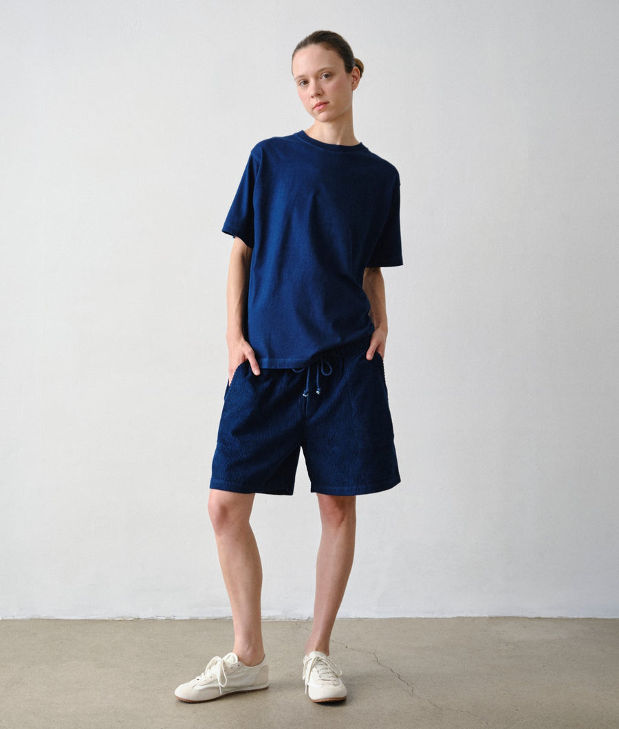A model stands against a plain light gray wall wearing IOAN Indigo 12 Ponya Corduroy Shorts, a matching T-shirt, and white sneakers.