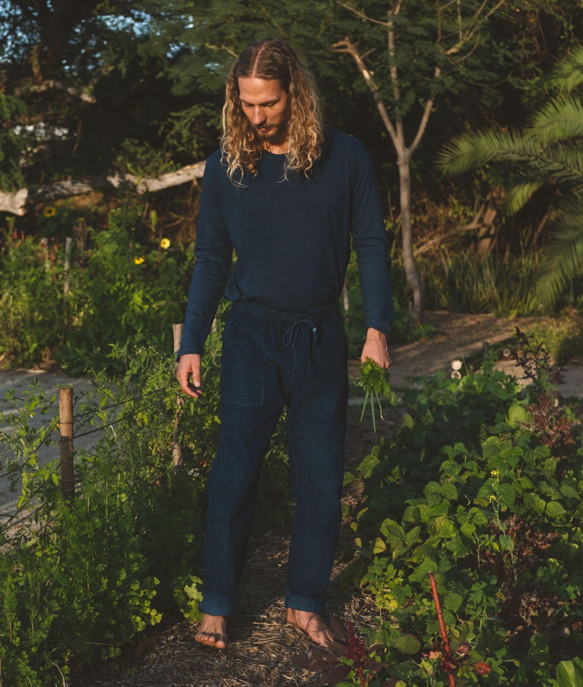 A man with long blonde hair wearing an IOAN Clean Crew Long Sleeve T-Shirt in Indigo 12 with matching corduroy pants walks through a lush green garden at sunset.