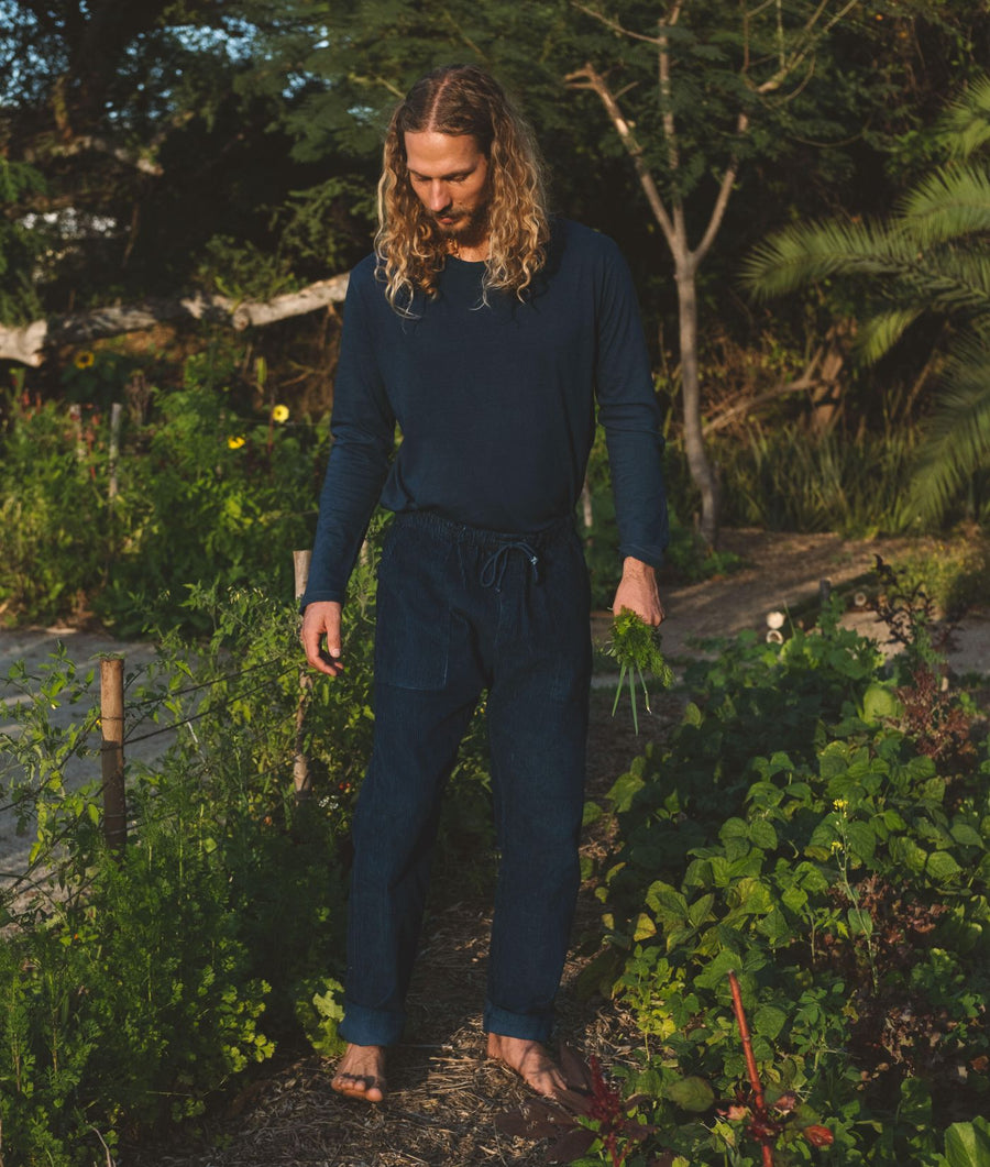 A man with long blonde hair wearing an IOAN Clean Crew Long Sleeve T-Shirt in Indigo 12 with matching corduroy pants walks through a lush green garden at sunset.