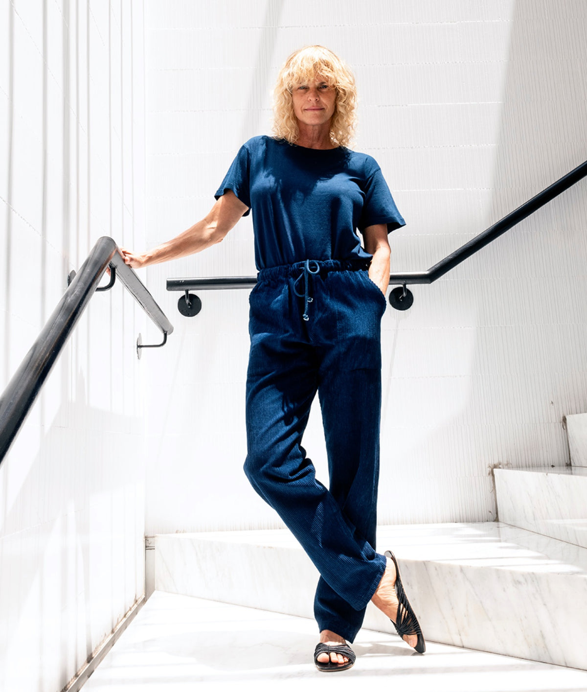 A blonde woman wearing an IOAN Clean Crewneck T-shirt and Ponya Drawstring Pants in Indigo 12 walks down white marble stairs against a white paneled wall.