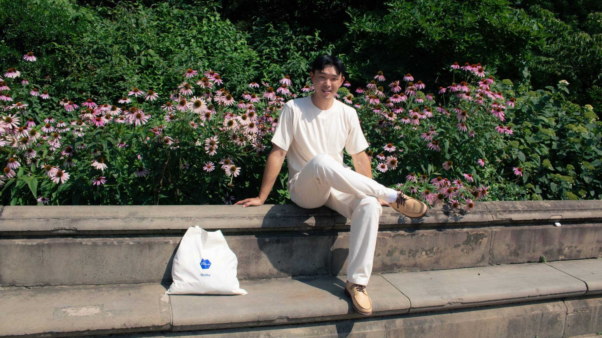 A man wearing an IOAN undyed T-shirt and matching Ponya Drawstring Pants sits on outdoor stone steps in front of a lush green garden with pink flowers. A white tote bag with a blue logo rests beside him.