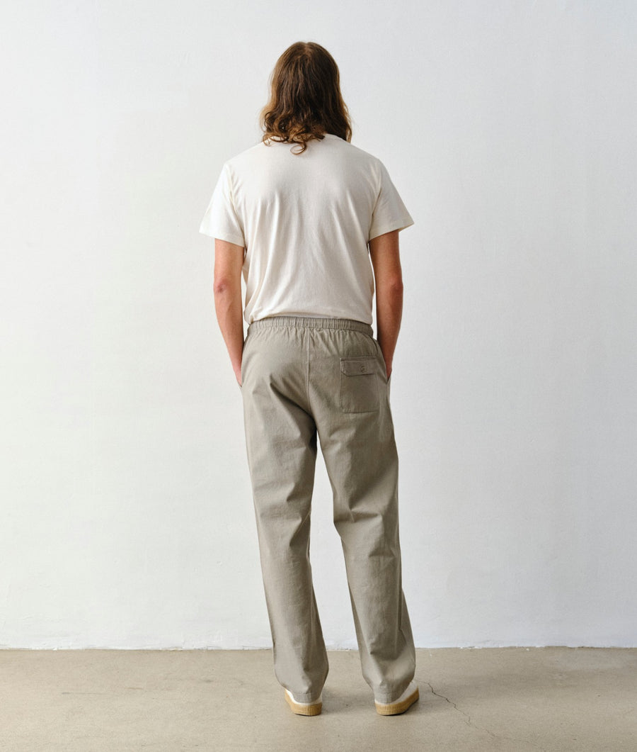 Man standing with his back to the camera, wearing an IOAN undyed T-shirt and Ponya Drawstring Pants in Iron 4 against a plain white wall.