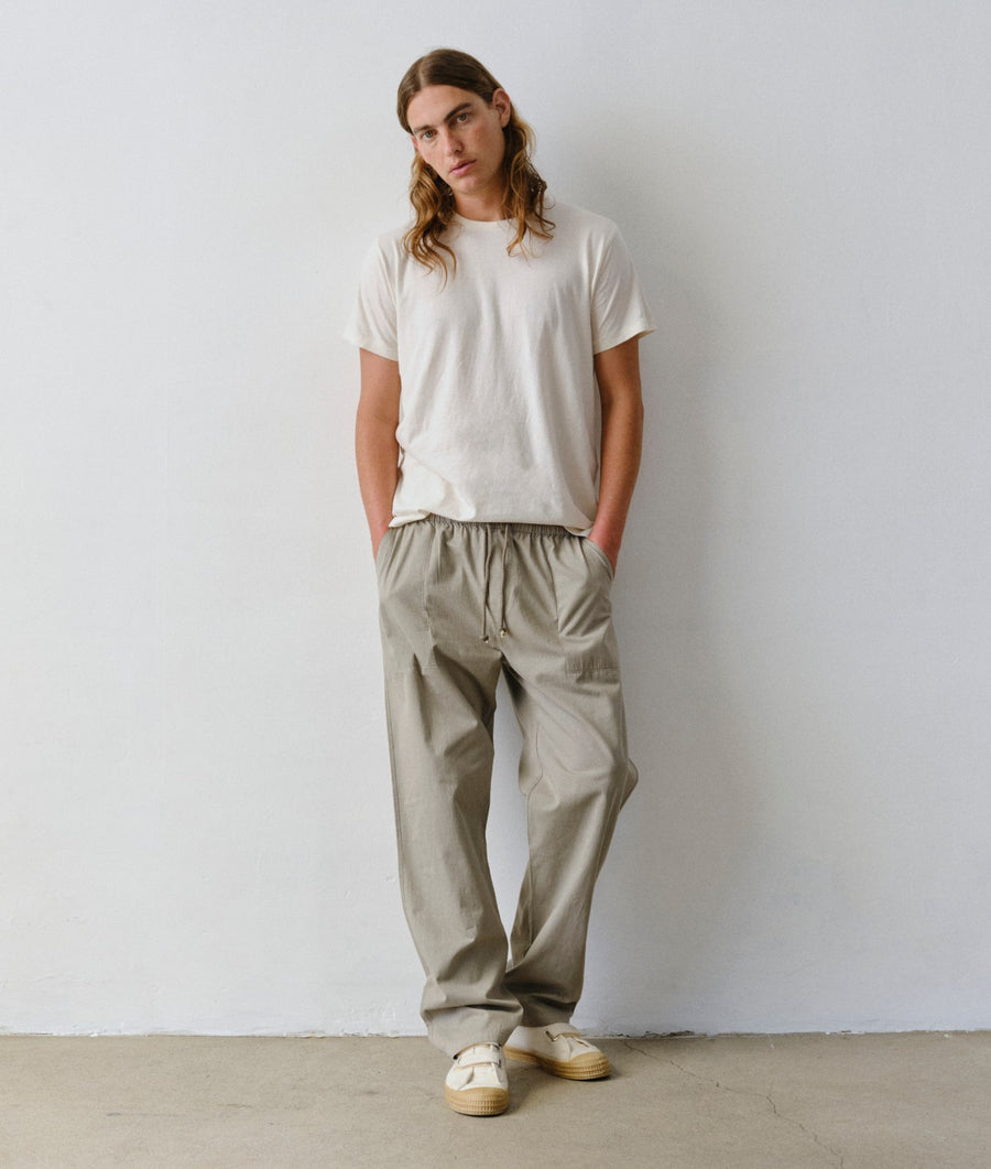 Man standing facing forward, wearing an IOAN undyed T-shirt and Ponya Drawstring Pants in Iron 4 against a plain white wall.