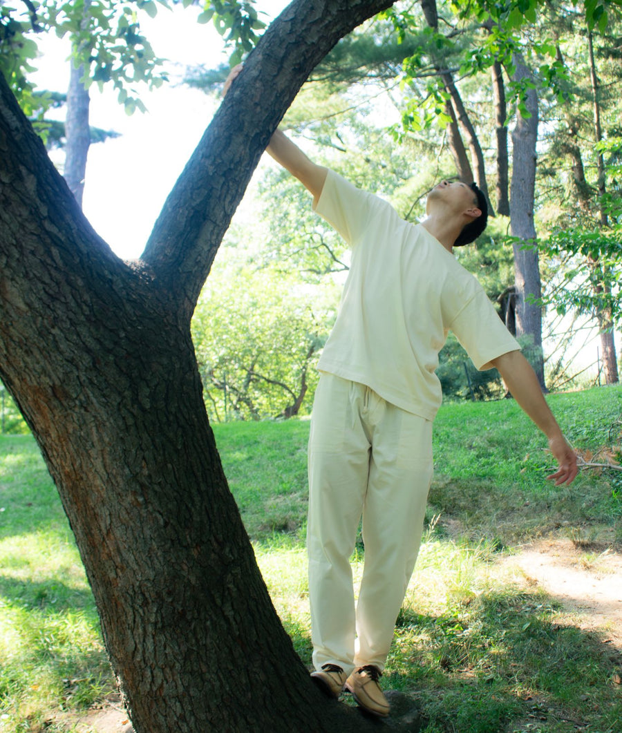 A man wearing the IOAN Ponya Drawstring Pants in Undyed and a matching cream-colored shirt stands under a tree in a grassy park, with his arms outstretched and looking upward.