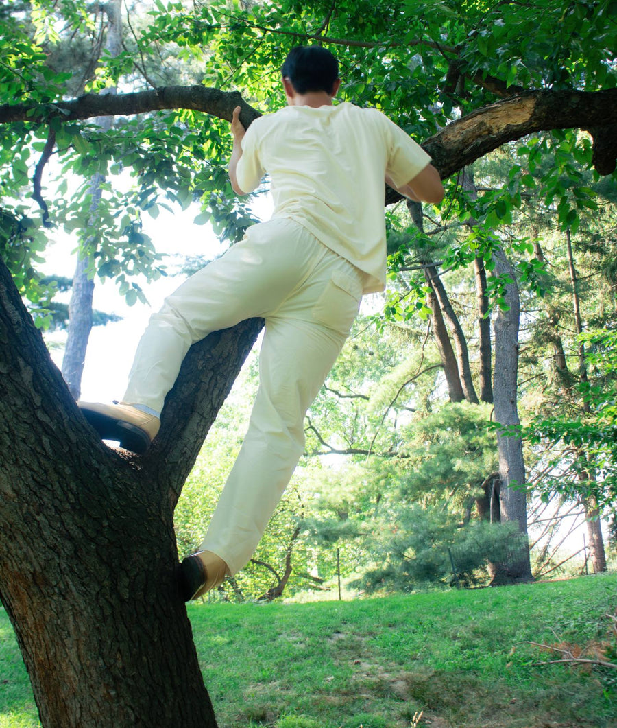 A man wearing the IOAN Ponya Drawstring Pants in Undyed and a matching cream-colored shirt climbs a tree outdoors in a grassy park setting.