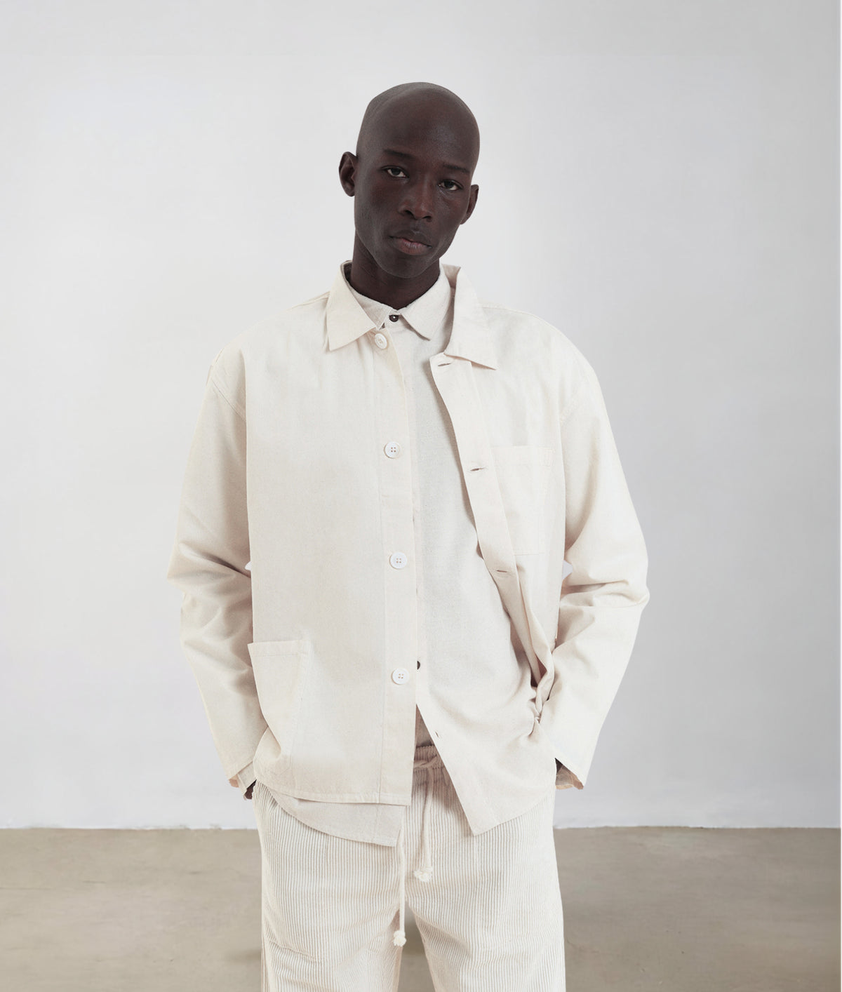 A man wearing an IOAN Ponya Jacket in Undyed with matching trousers, standing against a neutral light gray studio background.