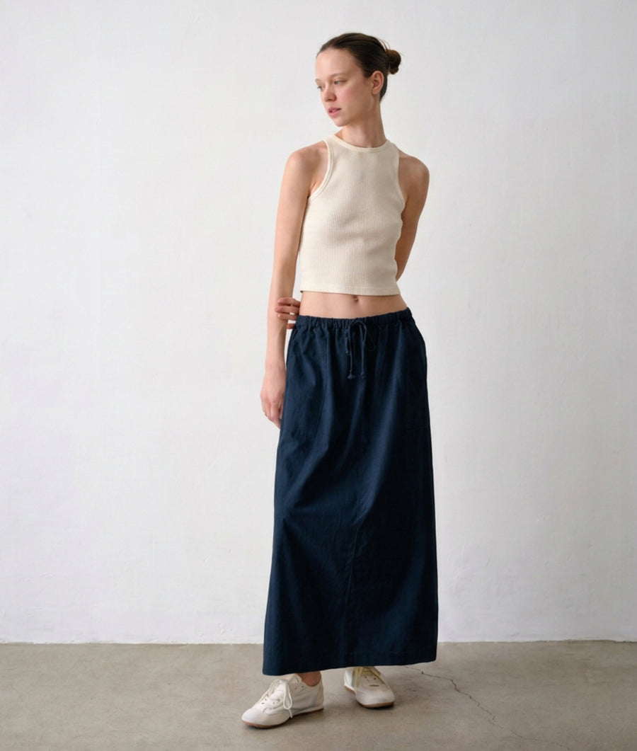 A woman stands in a minimalist studio wearing the IOAN Undyed Rib Tank, the IOAN Ponya Maxi Skirt in Indigo 12 with a drawstring waist, and white sneakers.