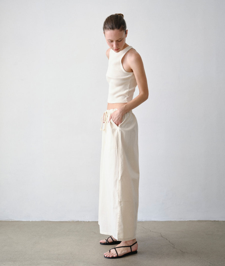 A model shown from the side in a minimalist studio setting wearing the IOAN Ponya Maxi Skirt in undyed and a matching IOAN rib tank.