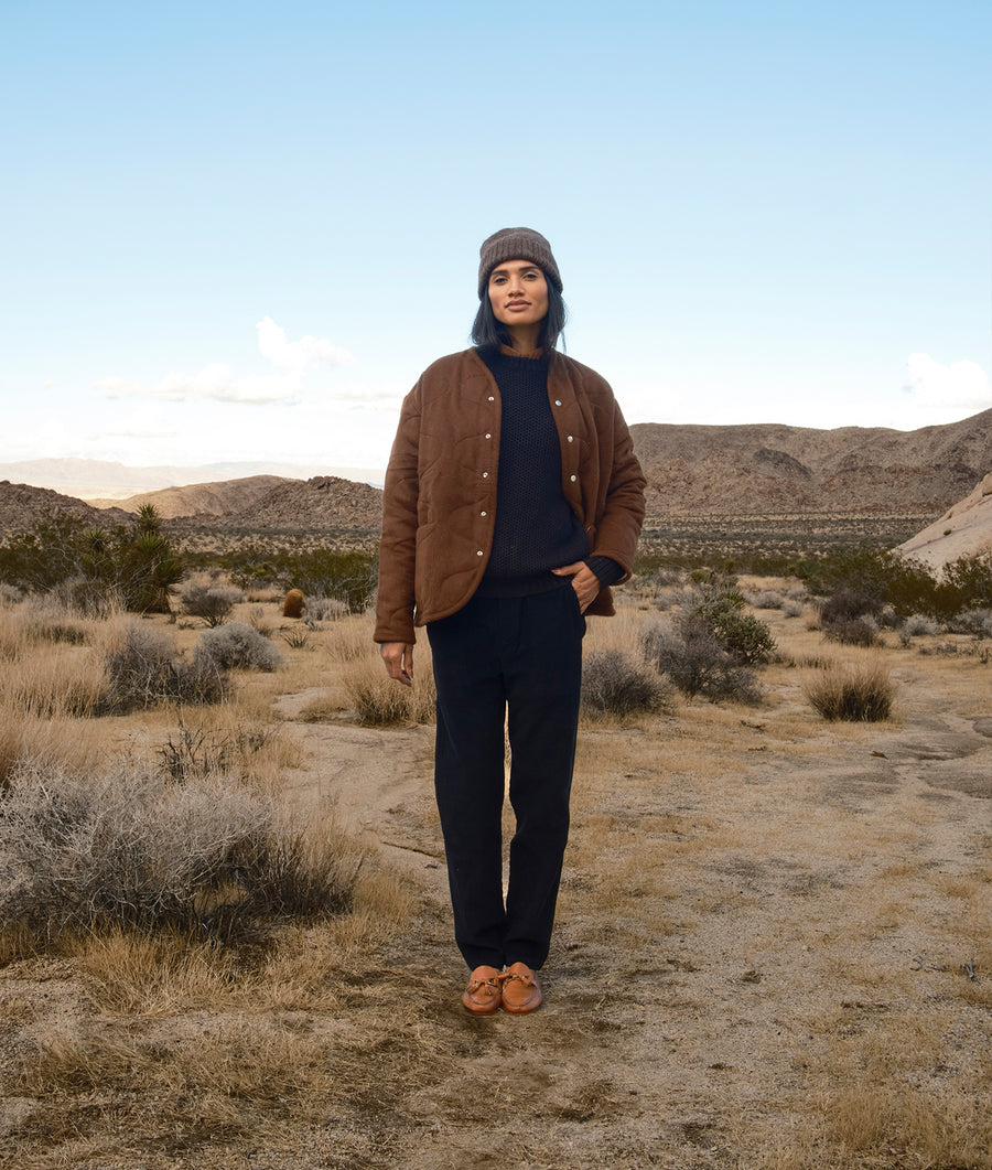 A person wearing a brown button-up jacket, standing in a desert landscape with mountains in the background - Industry of All Nations