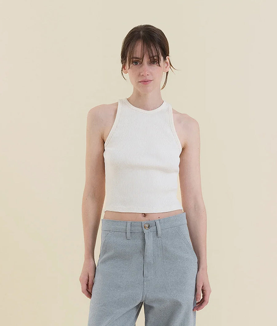 A woman wearing a white ribbed tank top and gray trousers, standing against a beige background - Industry of All Nations