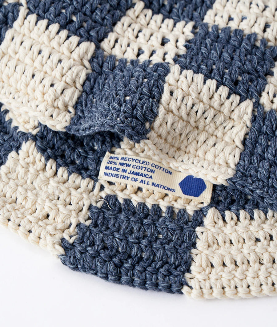 Detail shot of a crocheted blue and off-white circular rug or placemat with a tag reading "80% recycled cotton, 20% new cotton, made in Jamaica, industry of all nations" - Industry of All Nations