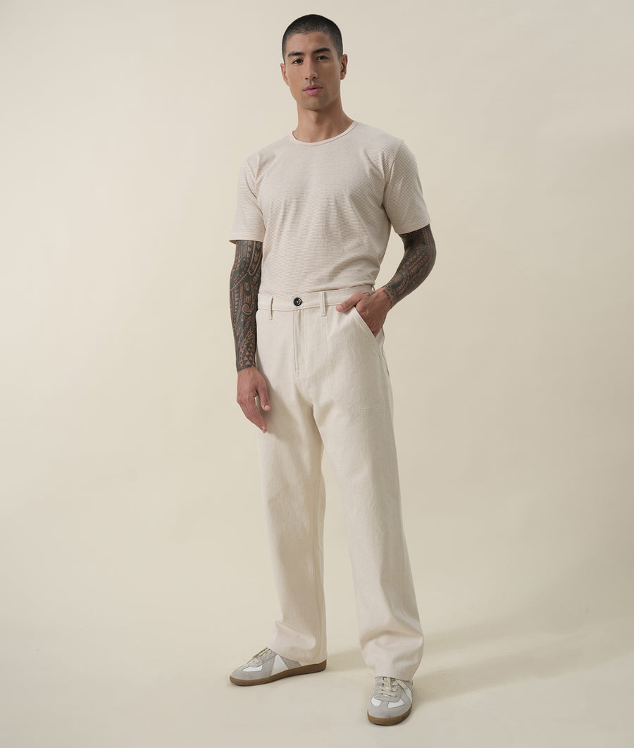 A man wearing a beige t-shirt and matching wide-leg pants, standing with hands in pockets - Industry of All Nations