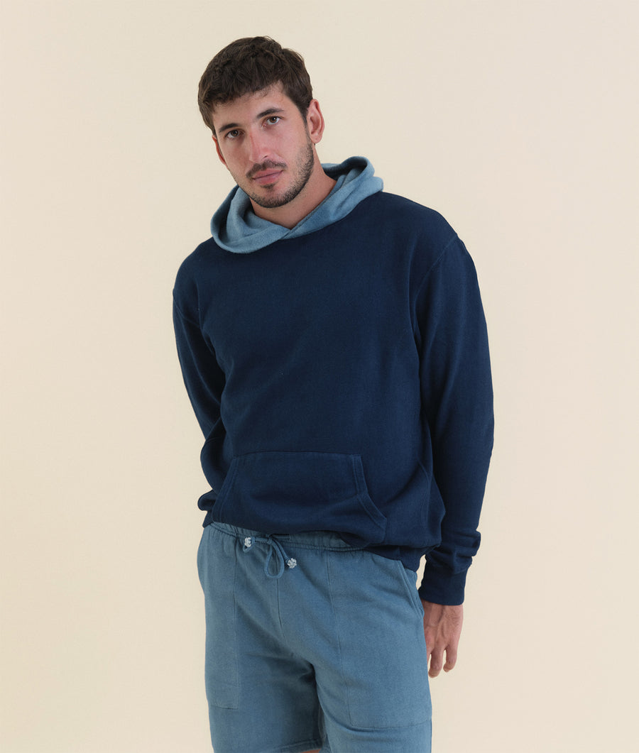 A man wearing a navy blue hooded sweatshirt with a light blue scarf, standing against a beige background - Industry of All Nations