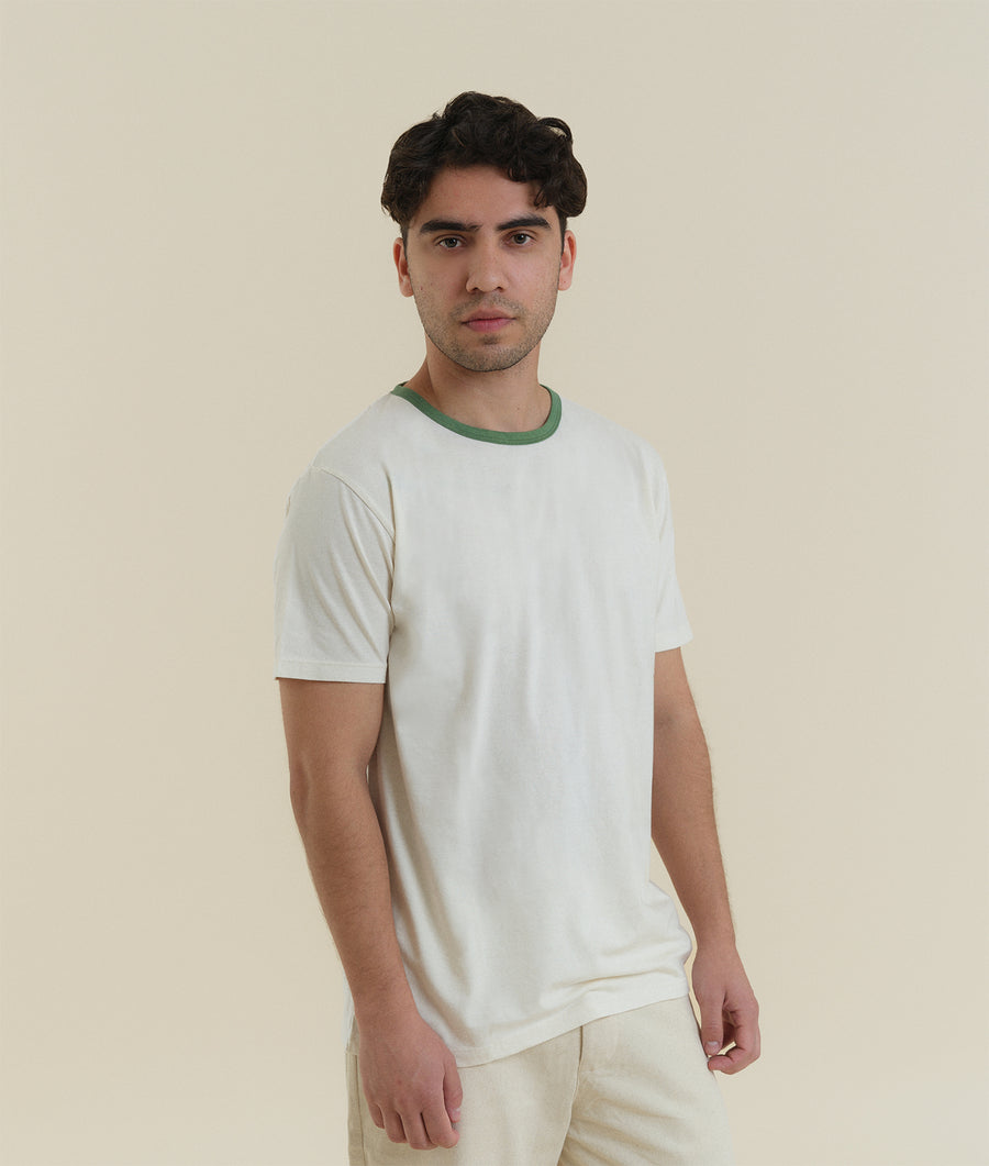 A young man wearing a white t-shirt with a green trim, posed against a neutral beige background - Industry of All Nations