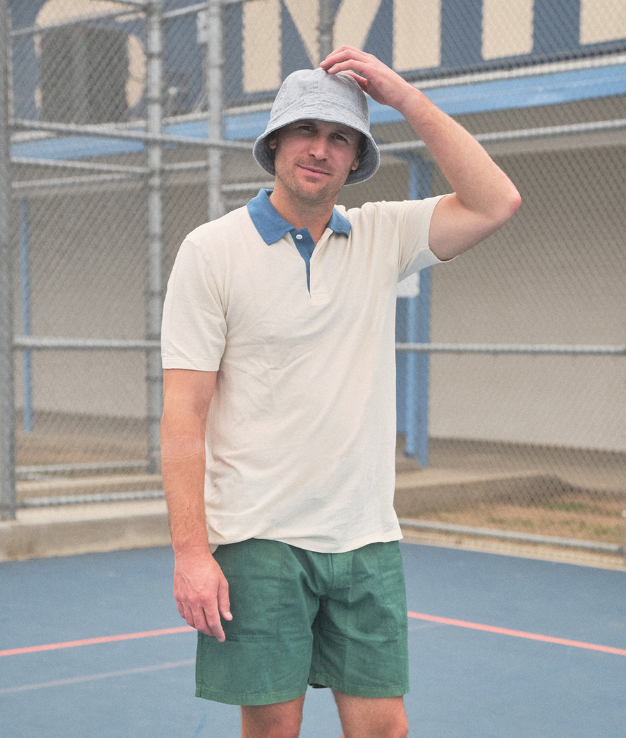 A man wearing a white polo shirt and green shorts standing outdoors against a wire mesh fence - Industry of All Nations
