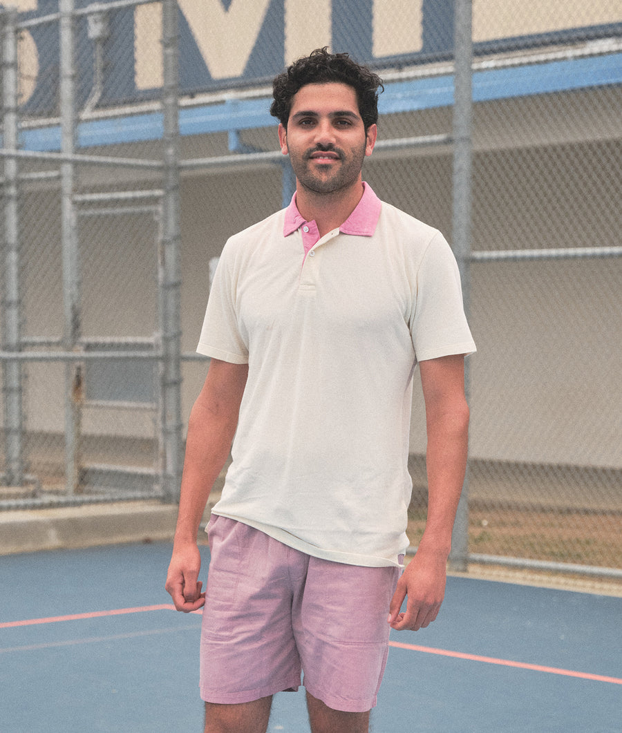 A man wearing a white polo shirt and light pink shorts stands in front of a blue tennis court fence - Industry of All Nations