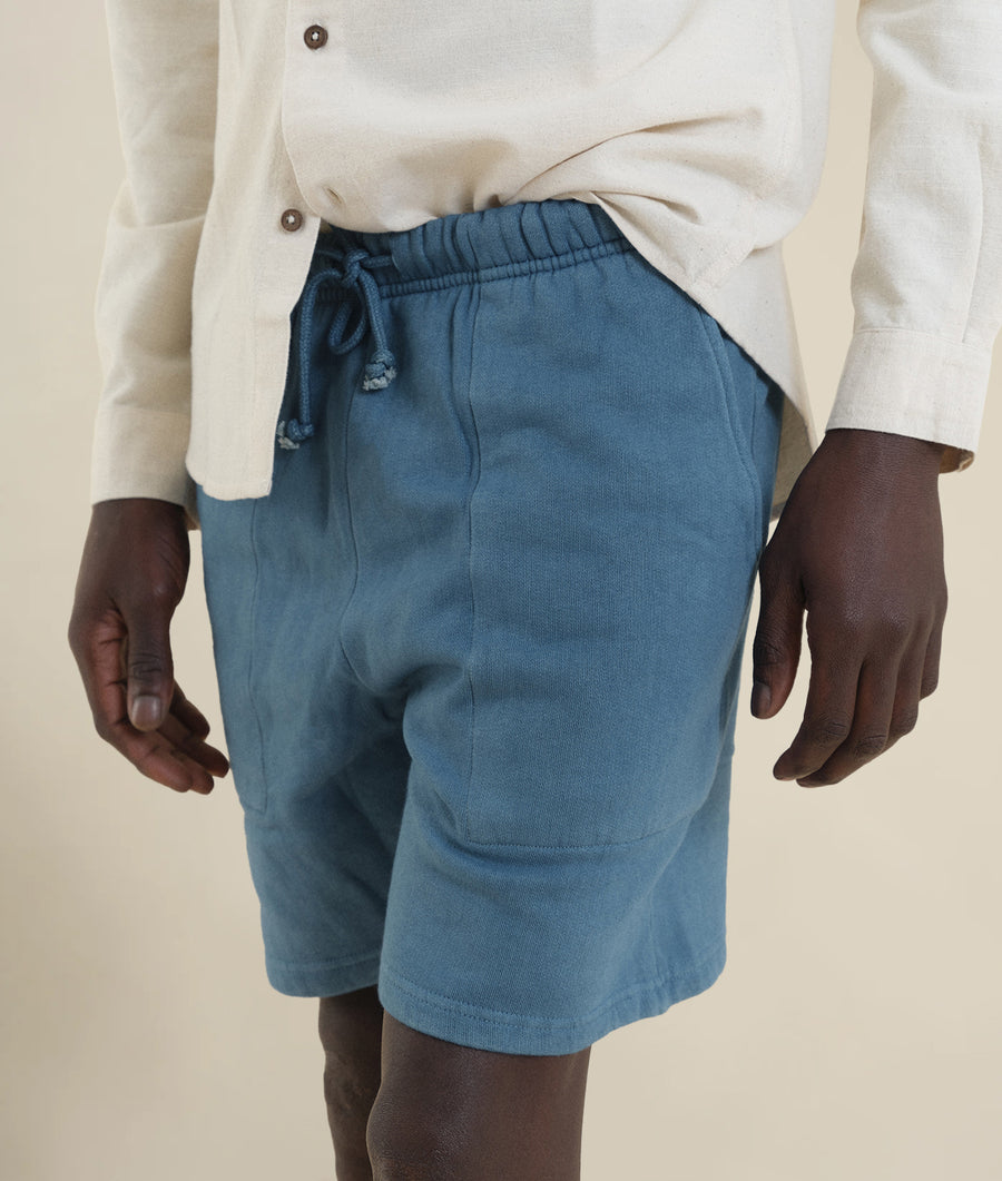 Image of a person wearing a pair of turquoise blue cotton shorts with an elasticated waist, displayed on a model wearing a white button-down shirt - Industry of All Nations