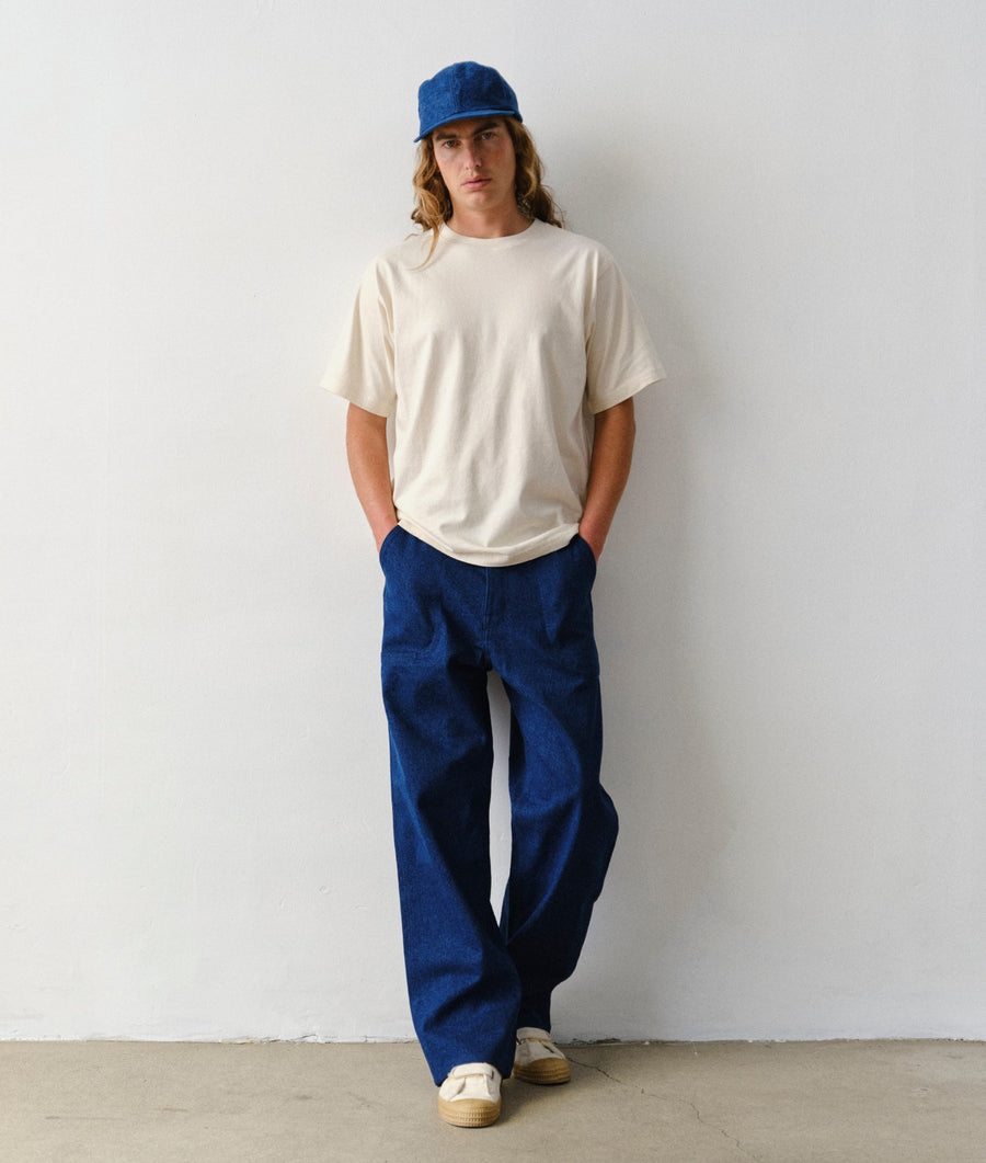 A man wearing IOAN Safari Pants in Azul and an undyed T-shirt, standing against a plain light gray wall.