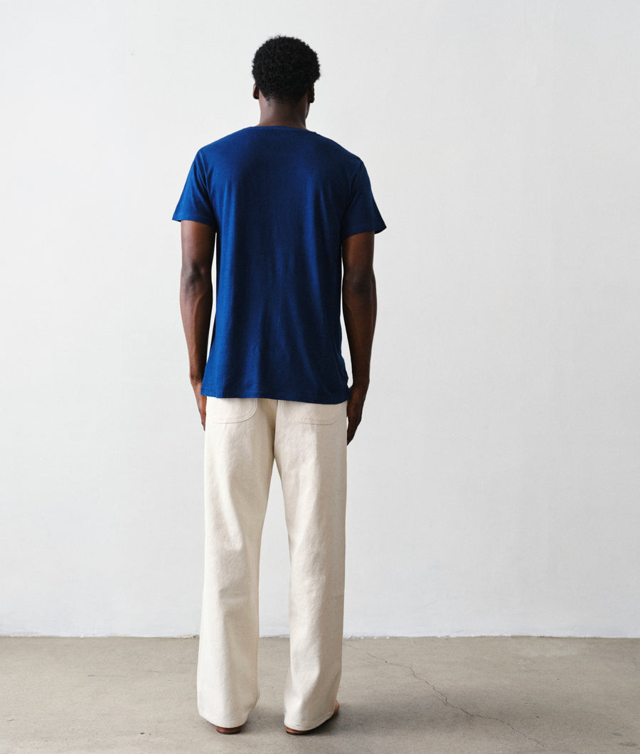 Back view of a man wearing an IOAN Clean Crewneck T-shirt in Indigo 12, undyed Safari Pants, and brown leather loafers, standing against a plain light gray wall.
