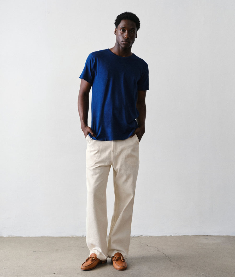 A man wearing an IOAN Clean Crewneck T-shirt in Indigo 12, undyed Safari Pants, and brown leather loafers, standing against a plain light gray wall.