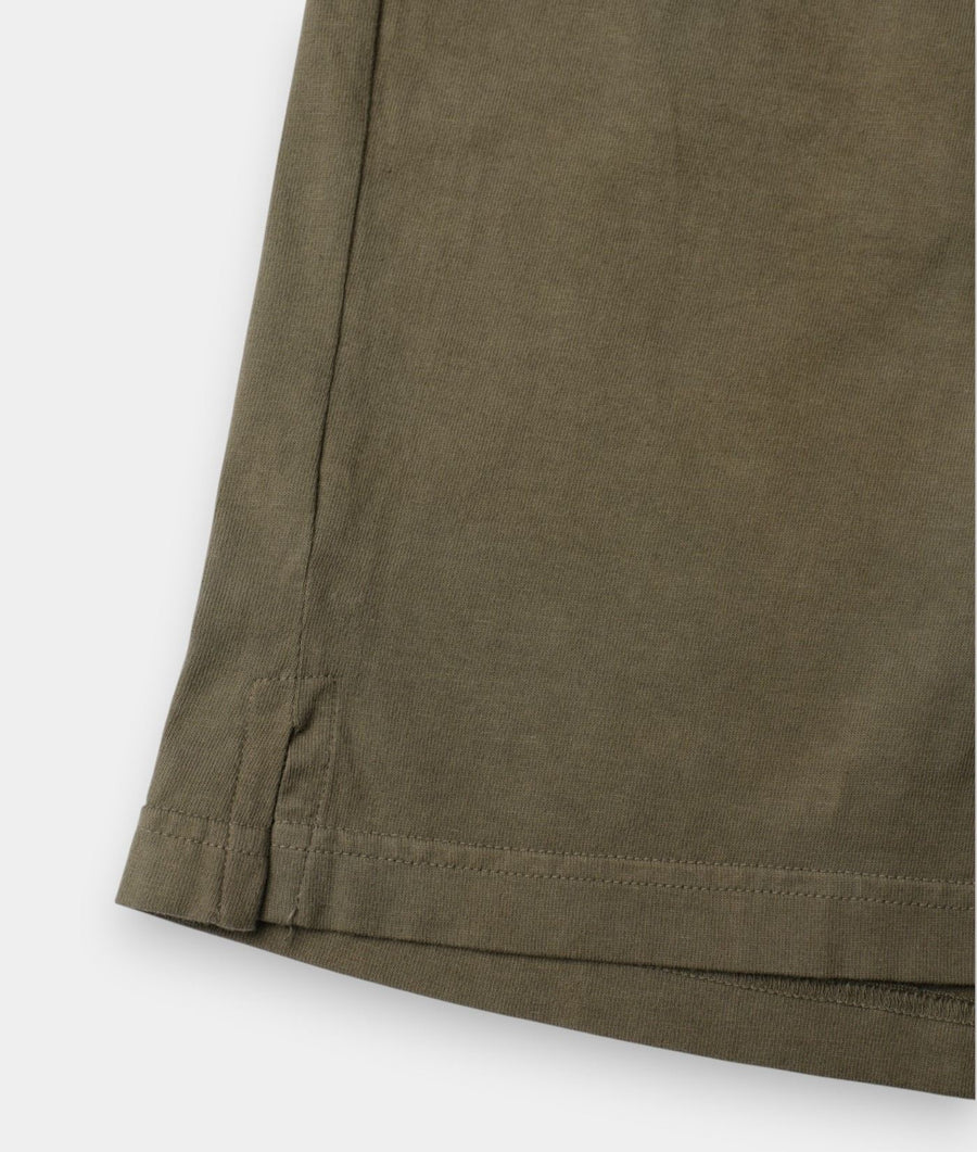 Close-up detail of an olive green garment, showing the hem with a stitched tab and clean stitching against a light gray background.