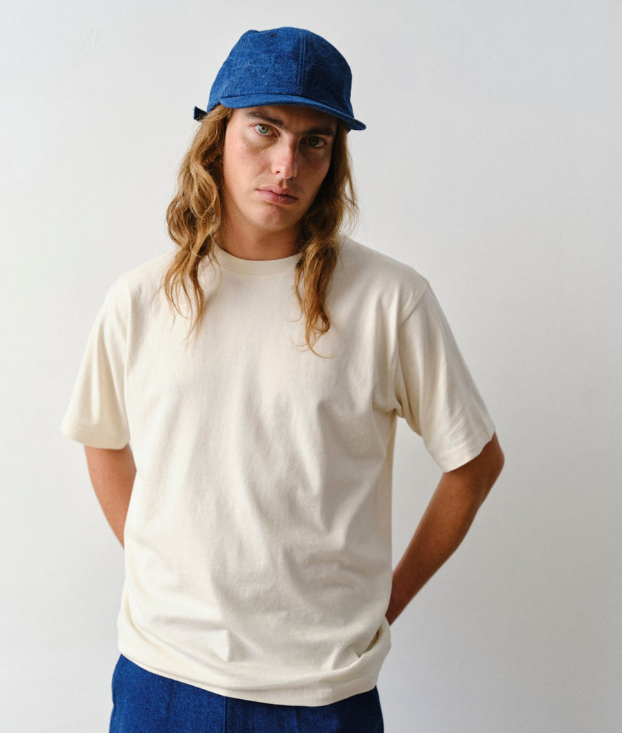 A man wearing the IOAN Undyed Standard T-Shirt, blue pants, and a blue cap, standing against a light gray background.
