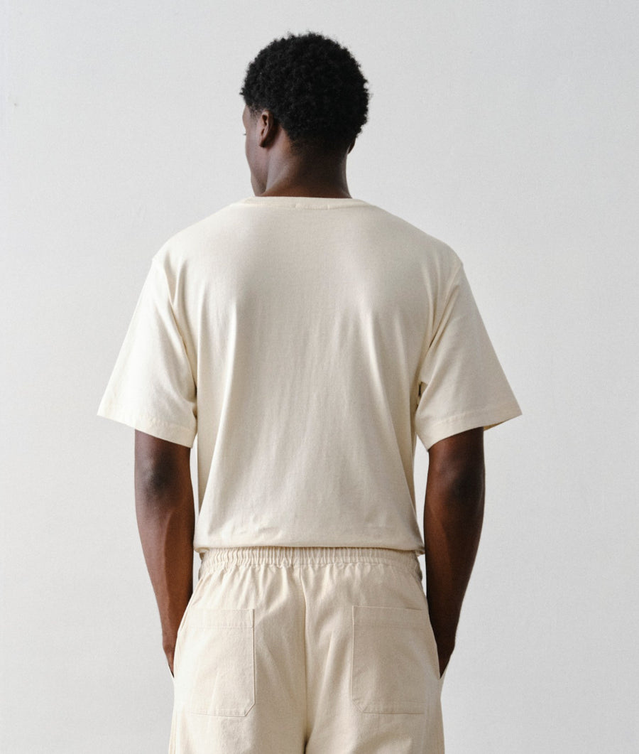A man viewed from behind wearing IOAN undyed  standard t-shirt and matching cream-colored pants against a plain light gray background