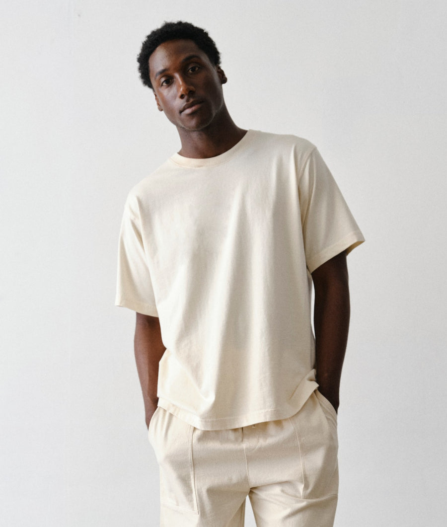 A man wearing IOAN undyed standard t-shirt and matching cream-colored pants, standing against a plain light gray background.