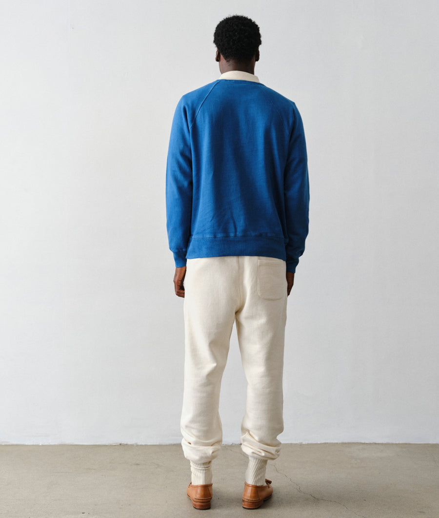 Back view of a man wearing IOAN Super Sweatpants in undyed natural color with a vibrant indigo blue IOAN Pocket Sweatshirt, white socks, and brown leather loafers, standing against a plain light gray wall.