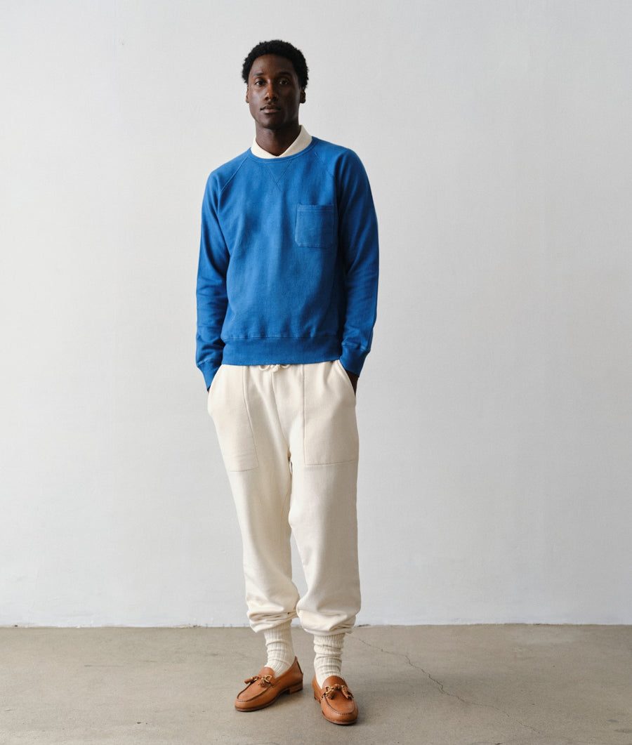 Man wearing IOAN Super Sweatpants in undyed natural color with a vibrant indigo blue IOAN Pocket Sweatshirt, white socks, and brown leather loafers, standing against a plain light gray wall.