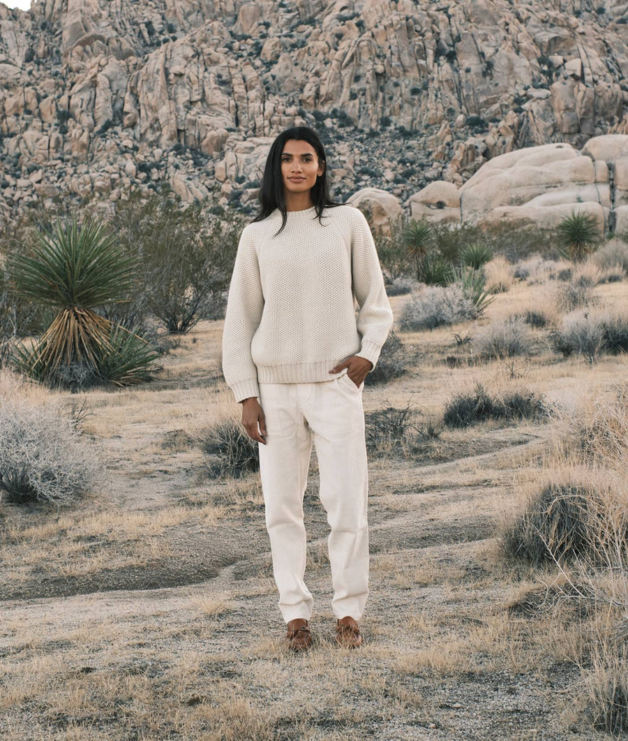 A woman in an ivory knit sweater and pants stands in front of a rocky desert landscape - Industry of All Nations