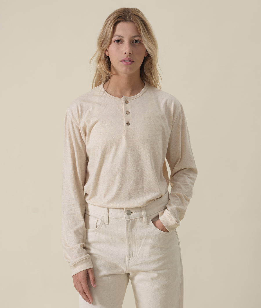 A young woman wearing a beige long-sleeved shirt with buttons down the front, standing with hands in pockets - Industry of All Nations