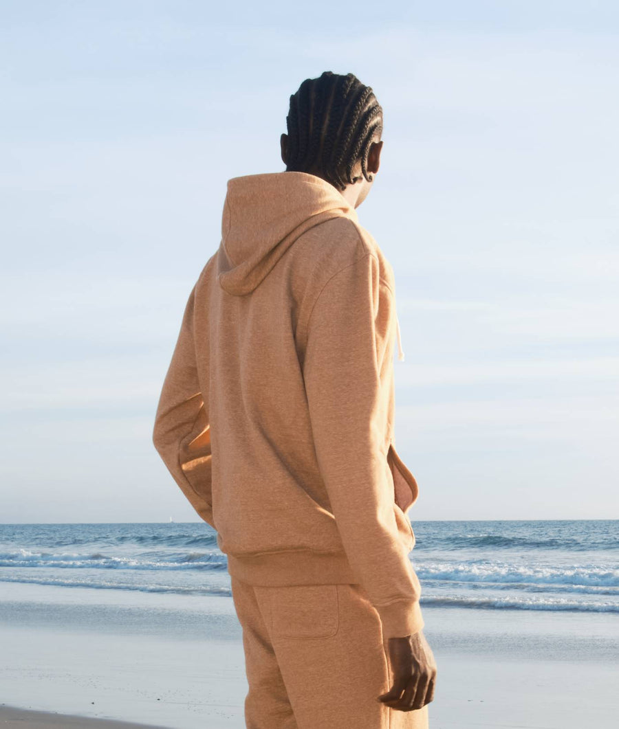 A person wearing a beige hooded sweatshirt while standing on a beach, facing the ocean - Industry of All Nations