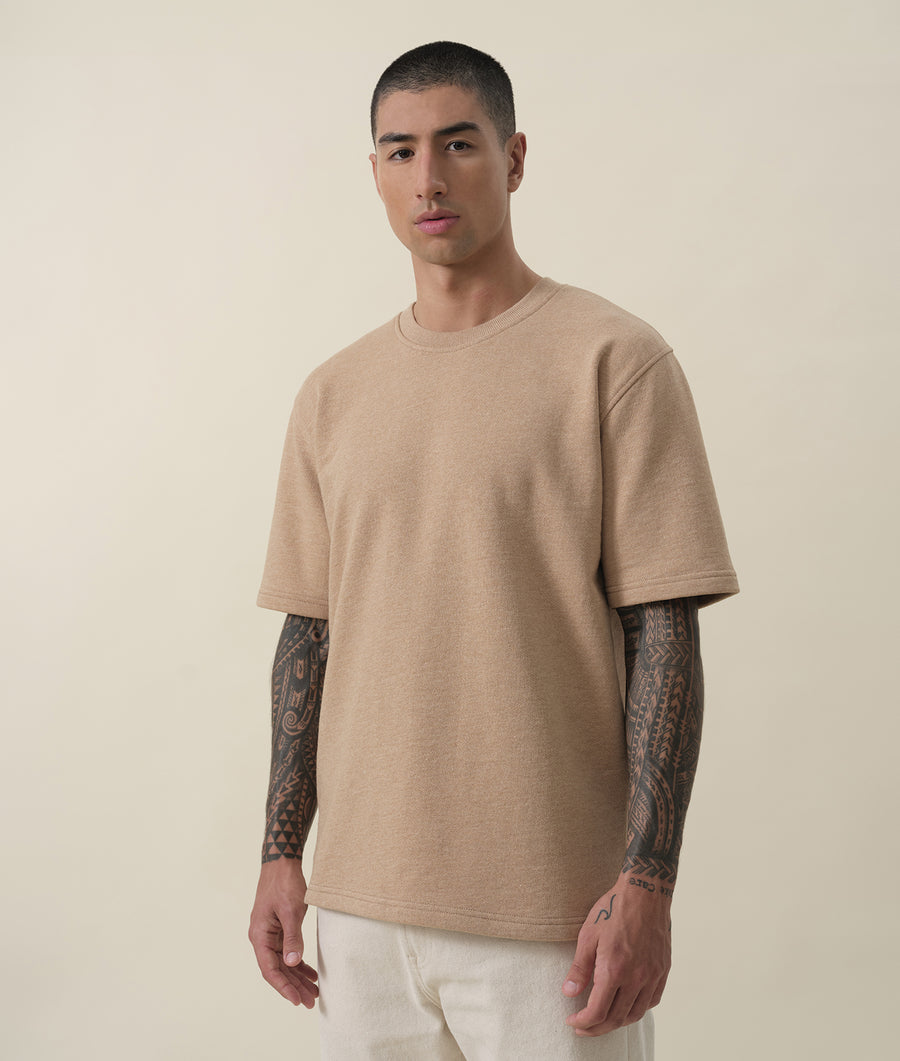 A man wearing a beige, crew neck t-shirt with a neutral expression on a plain background - Industry of All Nations