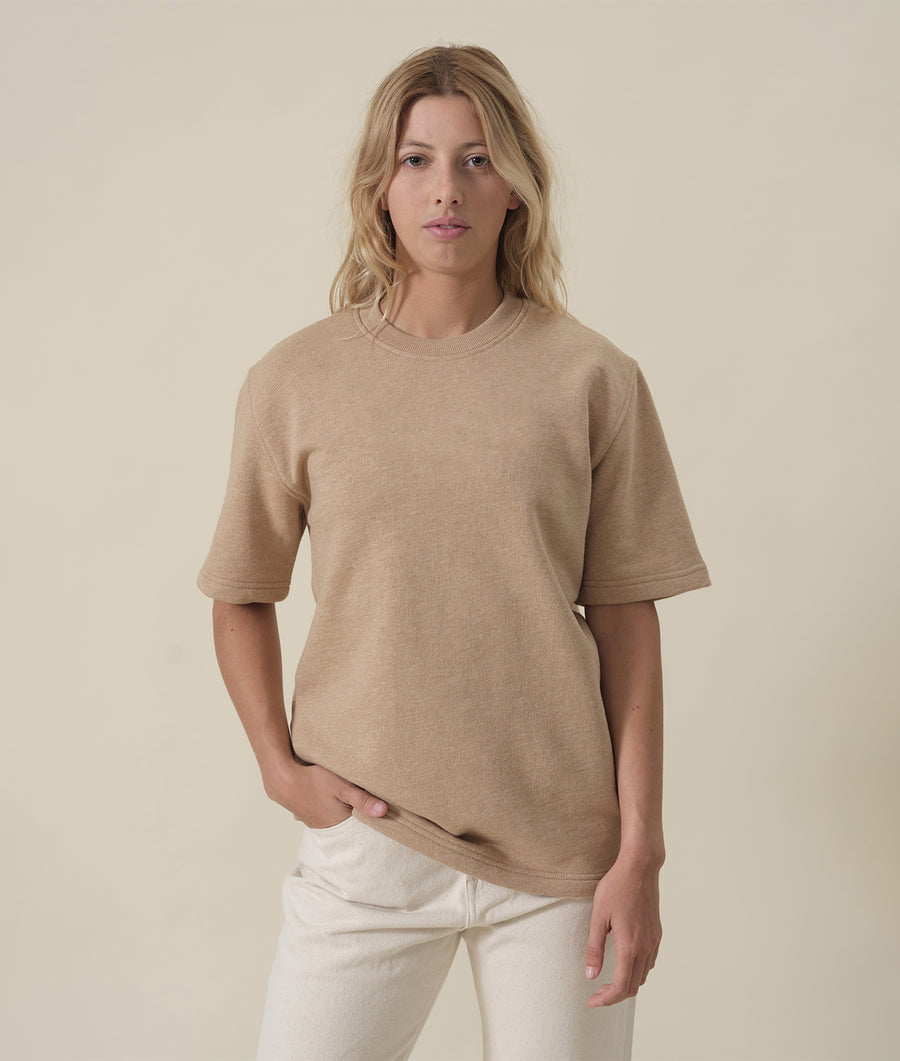The image shows a woman wearing a beige, ribbed-knit short-sleeve sweater from Industry of All Nations.