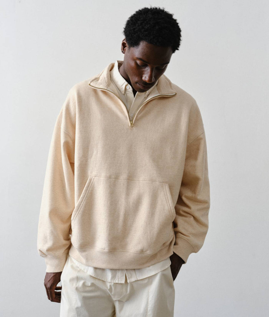 A man wearing a cream-colored quarter-zip pullover sweater and matching cream pants, standing against a plain light gray studio background.