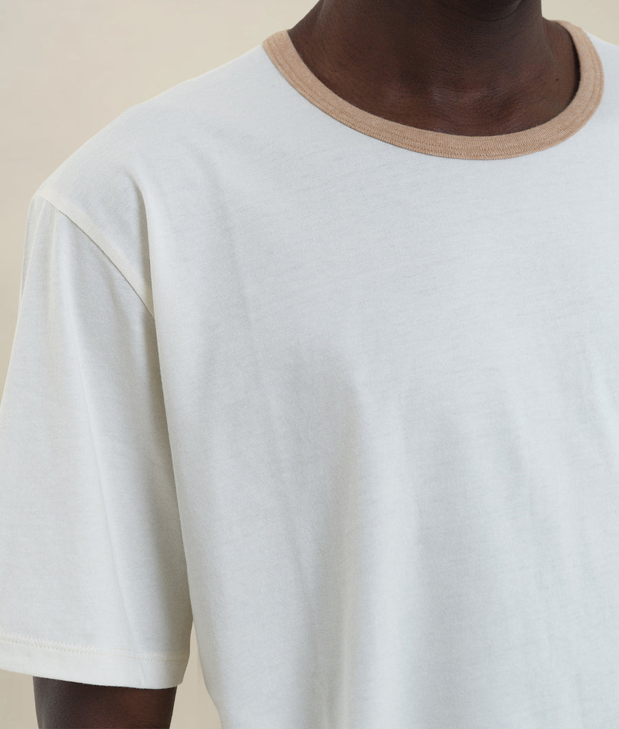 A close-up view of a white cotton t-shirt with a tan trim, worn by a model - Industry of All Nations