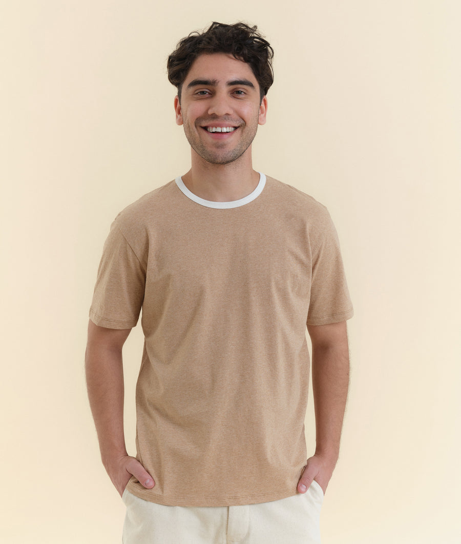 The image shows a smiling young man wearing a beige cotton t-shirt from Industry of All Nations.