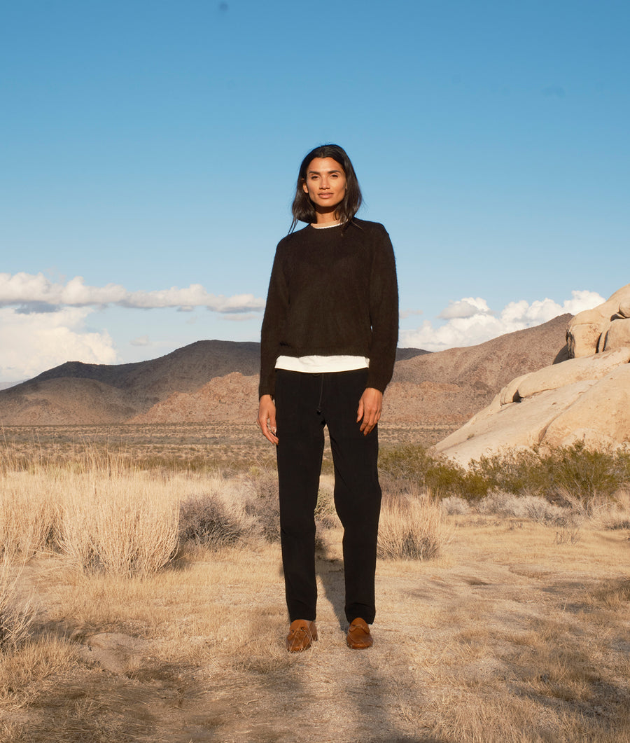 A woman wearing a black crewneck sweater and black pants stands in a desert landscape with mountains in the background - Industry of All Nations